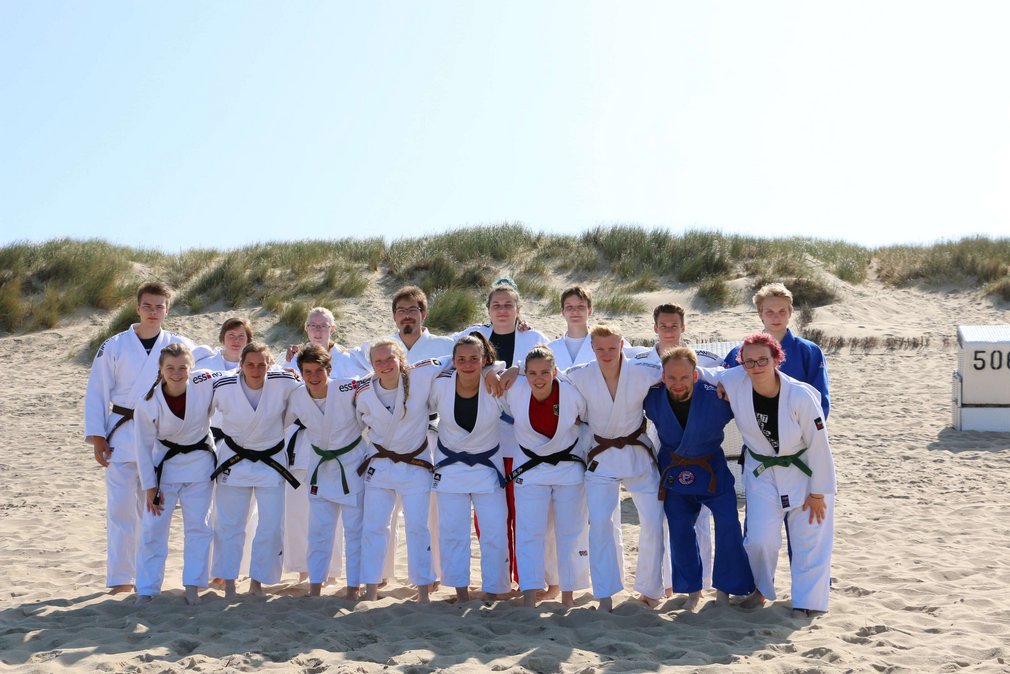 Eine Gruppe von Judoka posiert im Judoanzug fuer ein Gruppenfoto am Strand.
