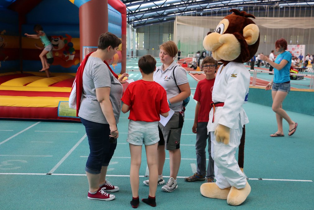 Momentaufnahme von einer Veranstaltung in einer Sporthalle. Eine Mitarbeiterin des DJB erklaert interessierten Personen etwas.