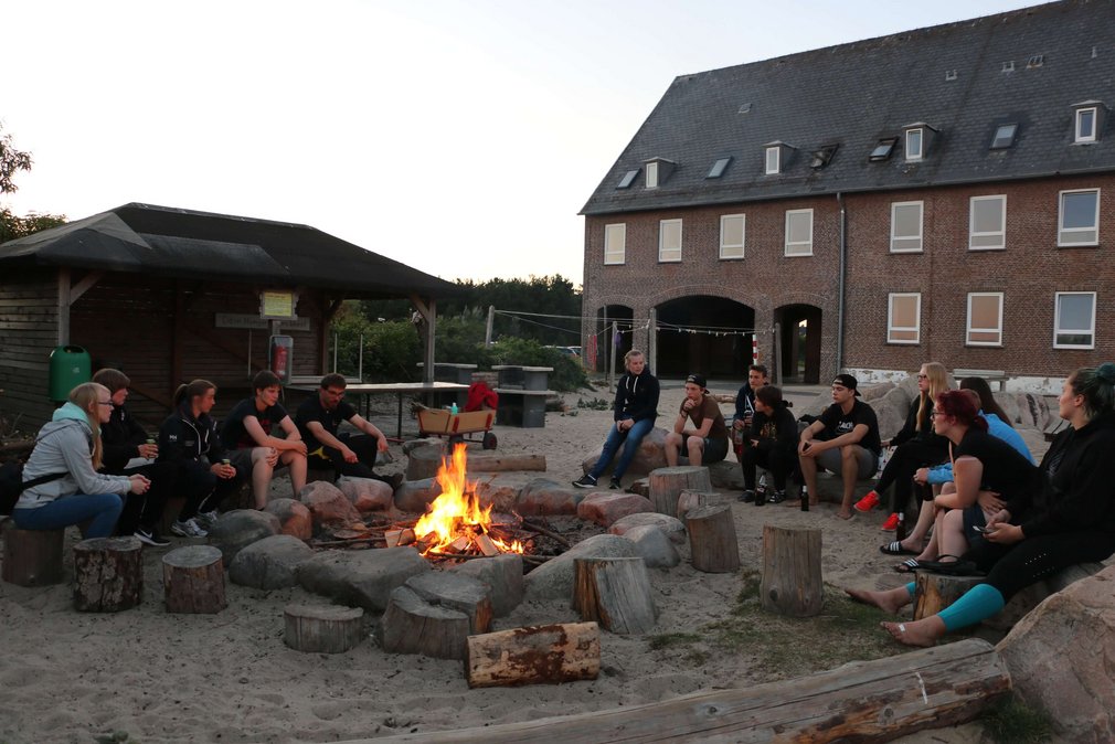 Eine Gruppe junger Menschen sitzt in der Daemmerung um ein Lagerfeuer.
