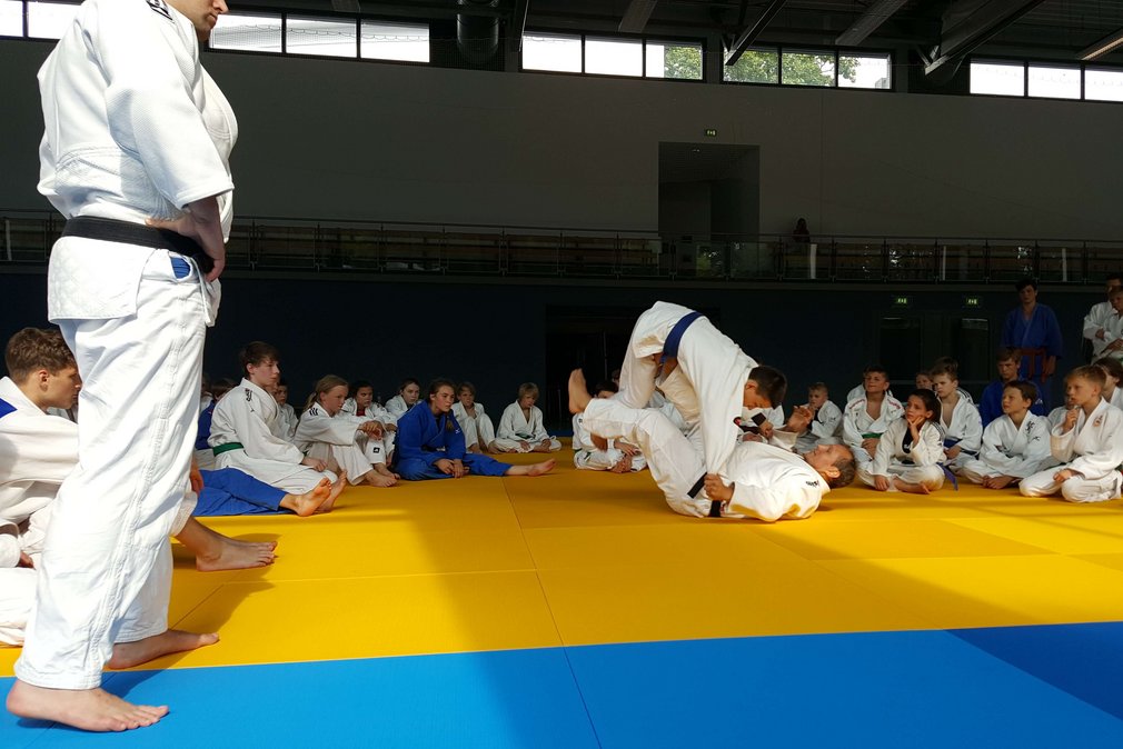 Eine Gruppe Judoka in einer Sporthalle. Zwei von ihnen kaempfen und die anderen schauen dabei zu.