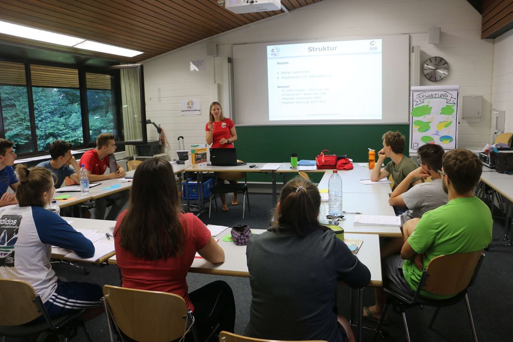Eine Gruppe junger Judoka sitzt in einem Klassenraum. Vorne steht eine Lehrperson und unterrichtet sie.