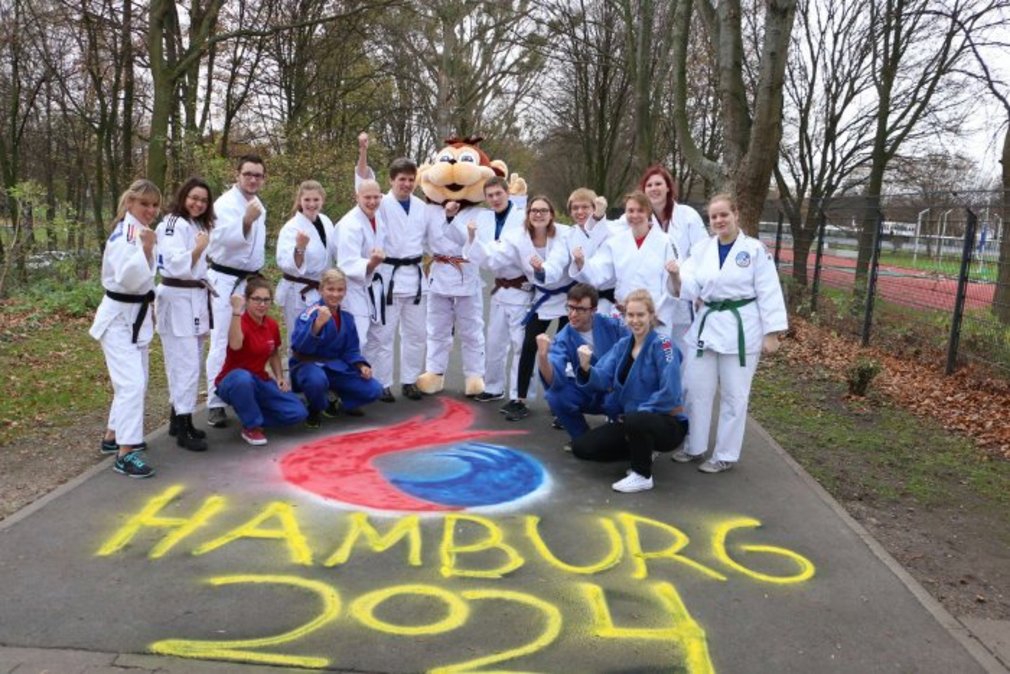 Eine Gruppe Judoka und Yoko posieren fuer ein Gruppenfoto auf einer Strasse, auf die HAMBURG 2024 gesprayt ist.