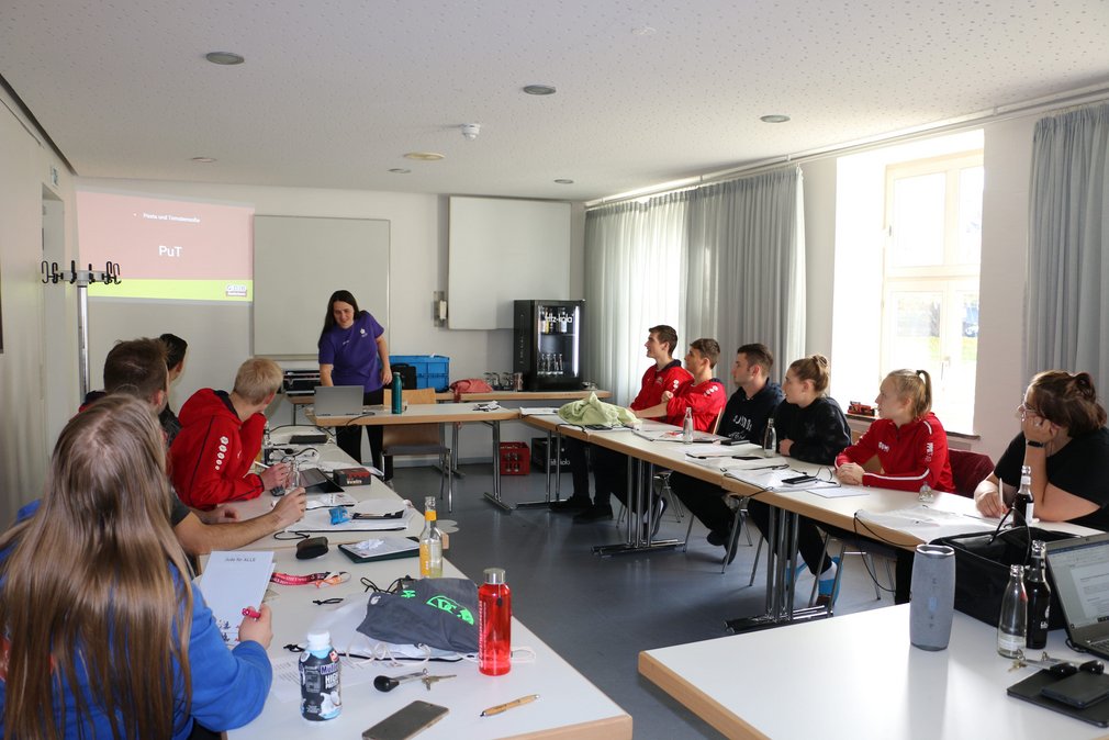 Eine Gruppe junger Menschen sitzt in einem Besprechungszimmer. Eine Person erklaert vorne etwas.