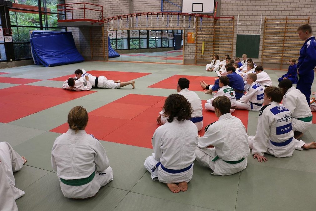 Zwei Judoka beim Trainingskampf waehrend andere im Kreis um sie herum sitzen und ihnen dabei zuschauen.