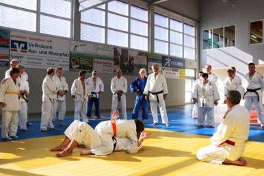 Zwei Judoka kaempfen am Boden und werden dabei von anderen Judoka beobachtet.