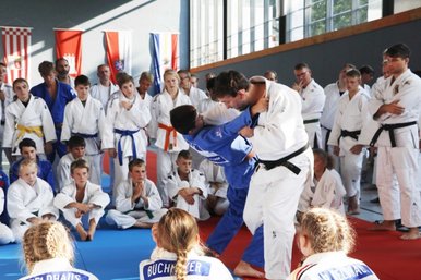 Viele Judoka stehen und sitzen im Kreis und schauen zwei Judoka beim Trainingskampf zu. 