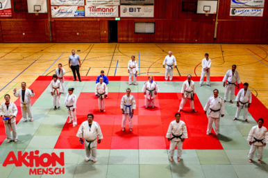 Judoka stehen verteilt auf einer Matte in einer Sporthalle. Alle sind der Kamera zugewandt.