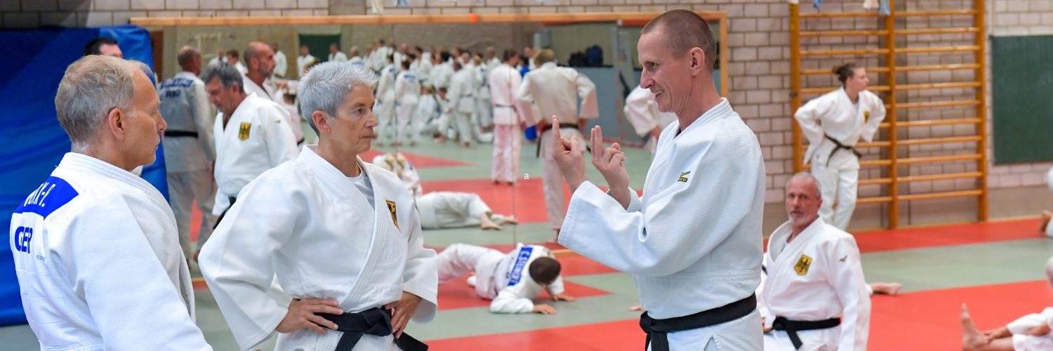 Drei Judoka im Gespraech. Im Hintergrund sind weitere Judoka verschiedenen Alters beim Training zu sehen.
