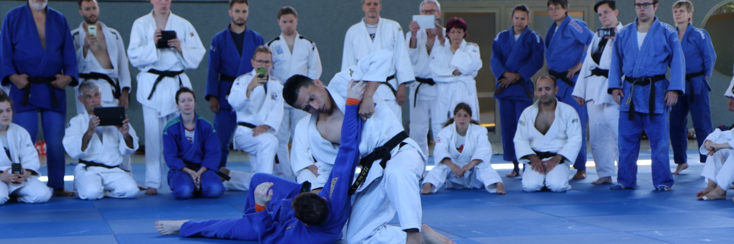 Zwei Judoka, die gegeneinander antreten. Im Hintergrund schauen weitere Judoka zu.