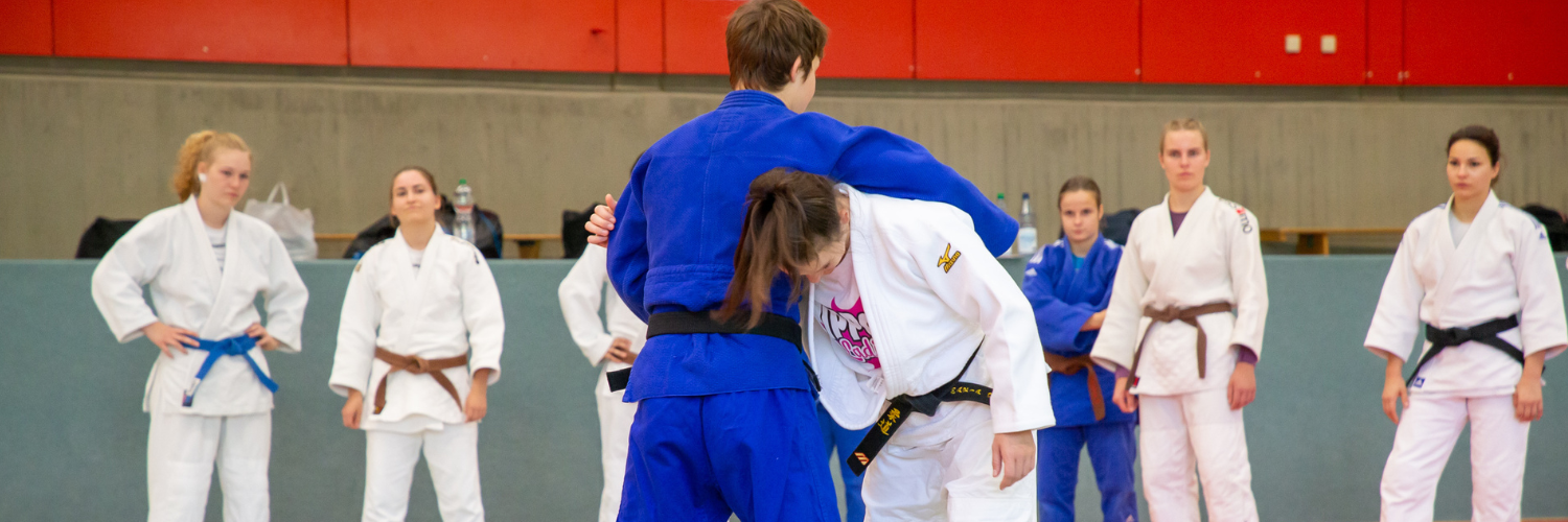 Zwei Judoka trainieren, waehrend weitere Judoka um sie herumstehen und zuschauen.