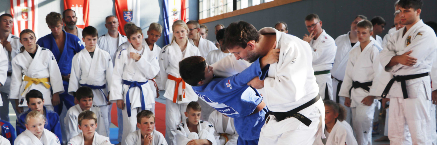 Zwei Judoka beim Trainingswettkampf und werden dabei von umstehenden Judoka beobachtet.