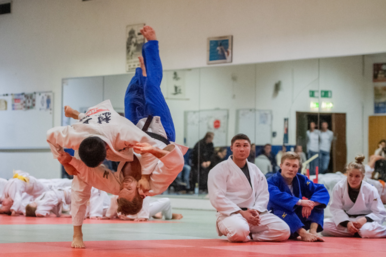 Zwei Judoka kaempfen gegeneinander. Weitere sitzen im Hintergrund und schauen ihnen zu.